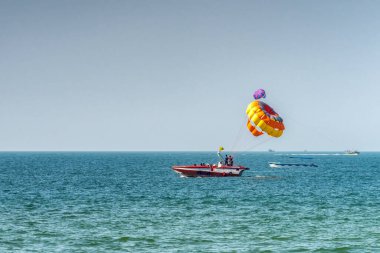 Tanınmayan insanlar teknede paraşütle atlamaya hazırlanıyorlar, yakında uçmaya başlıyorlar. Paraşüte odaklan. Tatil anlayışı, kumsalda ekstrem sporlar.
