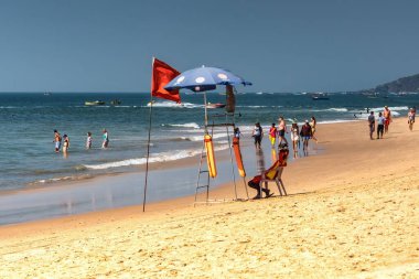 Ed CALANGUTE, GOA, INDIA HAZİRAN 3, 2019: Şemsiyenin altında oturan cankurtaran ve denize giden turistleri izleyen. Tehlike bayrağı, güneşli gün, bir sürü yürüyen turist.
