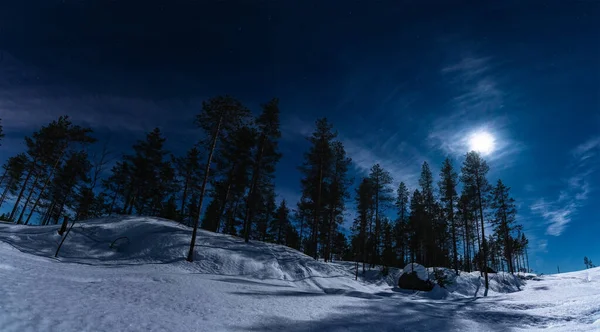 Full Moon lights over winter snowy forest with small hills, dark blue night sky with stars and some clouds highlighted by lunar light. Snowmobile trace, pine tree forest. Beautiful landscape