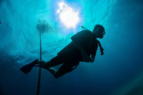 Silhouette of young diver against Sun light at the sea surface and hanging down steel anchor chain. Air bubbles from breathing. Scuba diving in tropical sea