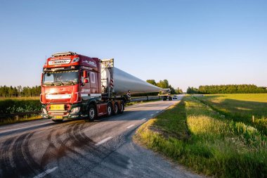 UMEA, SWEDEN - 4 Temmuz 2021: Küçük kırsal yolda çok uzun bir rüzgar türbini pervane bıçağının taşınması. Modern otoban traktörü, güvenlik aracı eşliğinde. Güneşli yaz akşamı.