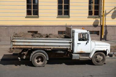 Şehir caddesinde park etmiş eski beyaz çöp kamyonu, kargo yatağı toprakla dolu, parlak güneş ışığı altında açık sarı bir binanın yanında duruyor güçlü gölgeler yaratıyor.