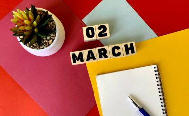 Tahta küplere 2 Mart. Yanında beyaz bir defter, bir kalem ve bir saksı çiçeği var. Mart ayı takvimi..