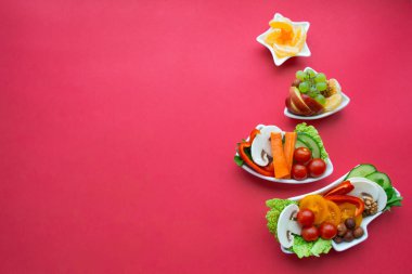 Tabak, içinde sebze, meyve, mantar ve böğürtlen olan bir Noel ağacı şeklinde. Kırmızı arka plan. Tatil için vejetaryen ziyafeti konsepti. Boşluğu kopyala.