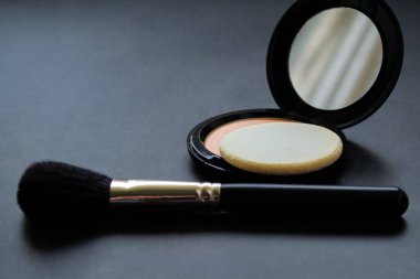 Aynalı, pudralı ve pembe tozlu açık bir kutunun yanında siyah bir makyaj fırçası. Siyah arka plan. Boşluğu kopyala.