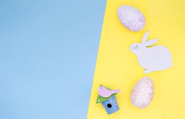 Paskalya Düz Yatağı: Paskalya Tavşanı, çiçekler, kuş yuvası ve sarı arka planda renkli yumurtalar, mavi arkaplanda yazı için boşluk.