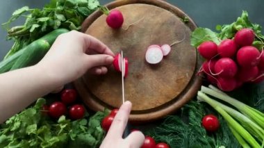 Elleri turpları beyaz seramik bir bıçakla kesiyor. Yuvarlak ahşap bir tahta üzerinde. Tahtanın etrafında olgun taze sebzeler var: domates, soğan, salatalık, kişniş ve dereotu.