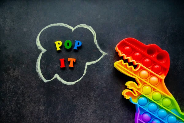 Pop it dinosaur toy rainbow colors on a black background with multicolored letters and the inscription - Pop it in a speech bubble.