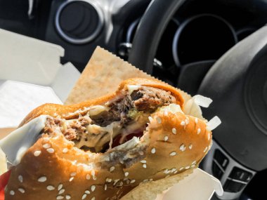 Araba sürerken yemek yemek: direksiyonun arka planına karşı arabanın içindeki sürücünün elinde ısırılmış bir hamburger..