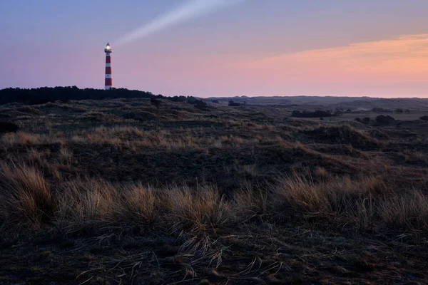 Hollum 'un tarihi deniz feneri, güneş doğarken kırmızı ve turuncu gökyüzü olan Ameland. Önünde sert çimenler olan karanlık kumullar.