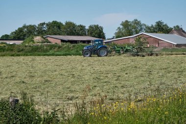 Dronten, Hollanda Haziran 12021: Büyük traktör biçme makinesi ve tırmığı olan otlak, çimenler fark edilmeye hazır. Arka planda bir çiftlik. Hollandalı çiftçiler saman biçmek, biçmek, tırmıklamak ve yetiştirmek için yağmura karşı yarışıyorlar.