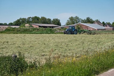 Dronten, Hollanda Haziran 12021: Büyük traktör biçme makinesi ve tırmığı olan otlak, çimenler fark edilmeye hazır. Arka planda bir çiftlik. Hollandalı çiftçiler saman biçmek, biçmek, tırmıklamak ve yetiştirmek için yağmura karşı yarışıyorlar.