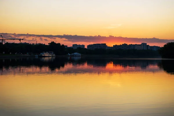 Sommersonnenuntergang am Ufer des Flusses. Blick auf die Stadt mit Spiegelungen im Wasser — Stockfoto