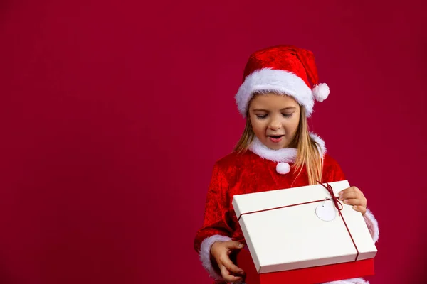 Noel Baba şapkası giyen sevimli küçük bir kız kırmızı arka planda boş bir alanda sürpriz bir hediye açıyor. Tatiller ve kış indirimi konsepti — Stok fotoğraf