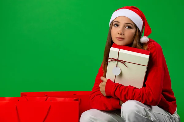 Симпатичная симпатичная девушка в шляпе Санта-Клауса обняла свой праздничный подарок возле зимних сумок для покупок и подумала. Концепция праздника, фото на зеленом фоне — стоковое фото