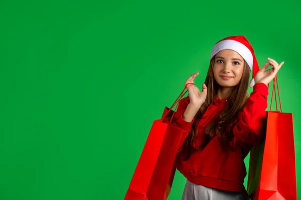 Yeşil arka planda poz veren güzel, mutlu bir kızın açık hava portresi. Model hediye çantaları tutuyor, kameraya bakıyor. Kış tatili, Noel indirimi, yeni yıl konsepti. — Stok fotoğraf