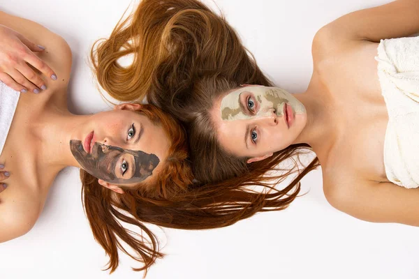 Two girls, blondine and redhaired, girlfriends with different masks on half face, lying on the floor wrapped in towels. Healthy skin and beauty concept on white background. High quality photo