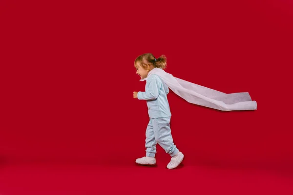 A little girl in blue pajamas, white slippers and a white cloak of a handmade hero runs and the cloak is opened. Concept on red background with free side space. High quality photo
