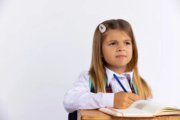 穿着校服的小女孩坐在白底的桌旁 空荡荡的 这个女学生正在做作业 若有所思地把目光移开 教育概念 高质量的照片 — 图库照片