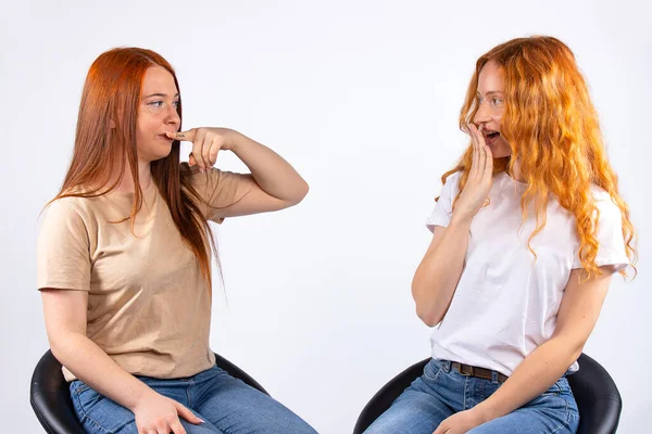 Two red-haired sisters sitting on leather chairs on a white background. Young girls fool around in front of the camera. They make faces and gesture: silence and wow. High quality photo