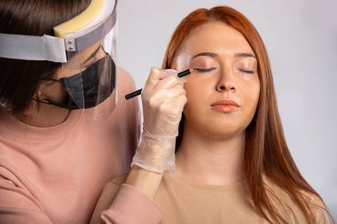 Uzun kızıl saçlı bir kız için zarif bir makyaj. Üstat makyaj malzemesi, maske, eldiven ve virüsten korunmak için koruyucu bir perde takıyor. Güzellik ve covid 19 konsepti. - Evet. Yüksek kalite fotoğraf