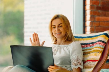  Hoş, sevimli bir kadın gülümsüyor, video konferans yapıyor ve el sıkışıyor. Arka planı bulanık, sallanan rahat bir sandalyede otururken selamlaşıyor. Yüksek kalite fotoğraf