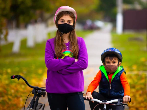 Брат і сестра в осінньому парку, їздять на велосипедах в захисних масках. На задньому плані розмитий осінній парк. Пандемія та концепція вірусу — стокове фото