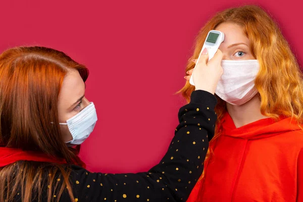 Duas meninas com máscaras médicas medem a temperatura corporal com um termômetro sem contato. Foto sobre um fundo vermelho. Conceito de Natal, vírus e pandemia — Fotografia de Stock