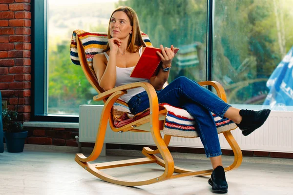 Genç ve çekici bir kadın kitap okuyor, sallanan rahat bir sandalyede sallanıyor. İyi vakit geçirmek seni neşelendirir.. — Stok fotoğraf