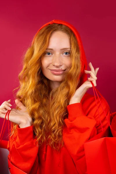Πορτρέτο ενός νεαρού κοριτσιού με κόκκινα μαλλιά και φακίδες που κρατάει κόκκινες σακούλες δώρων σε κόκκινο φόντο. Έννοια διακοπών και αγορών — Φωτογραφία Αρχείου