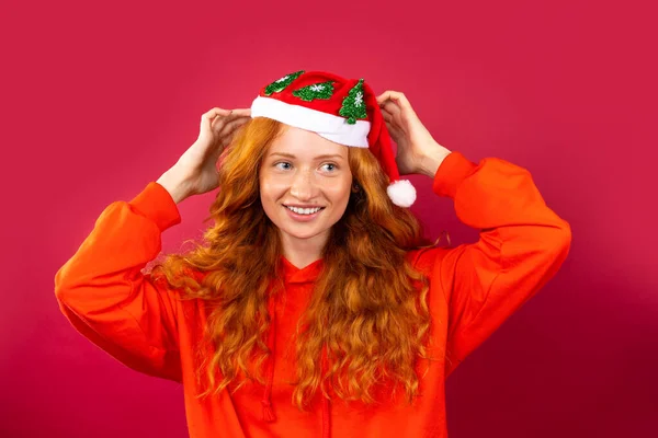 Chica pelirroja feliz, con una sonrisa brillante y hermoso pelo rizado, en un sombrero de Santa Claus sobre un fondo rojo. Concepto de vacaciones — Foto de Stock