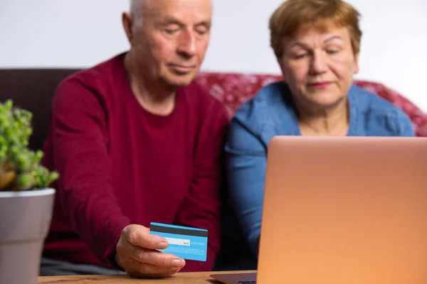 Ένας άντρας και μια γυναίκα σε ηλικία, με ένα λάπτοπ και μια πιστωτική κάρτα στο τραπέζι, κοντινό πλάνο. Πληρώστε για online παράδοση. Senior online και πανδημία έννοια — Φωτογραφία Αρχείου