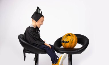 Yarasa kostümlü bir çocuk siyah sandalyelerde balkabağıyla oturuyor. Onu yanında görünce, mutlu bir şekilde gülümsüyor..