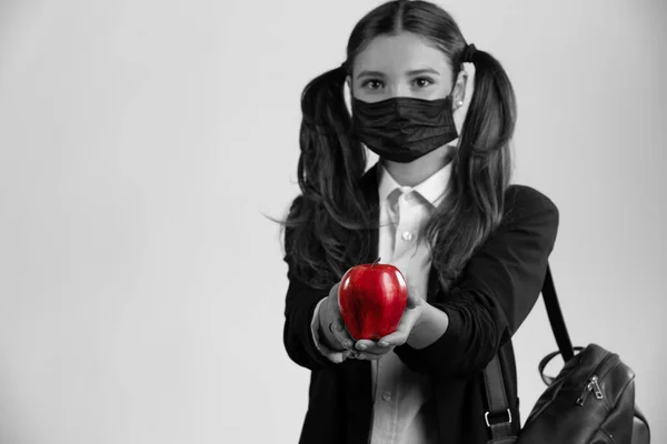 Schwarz-Weiß-Foto. Ein Studentenmädchen in Uniform mit Rucksack und medizinischer Maske hält einen roten Apfel in der Hand. Gesunde Ernährung und Vitamine gegen das Virus-Konzept — Stockfoto