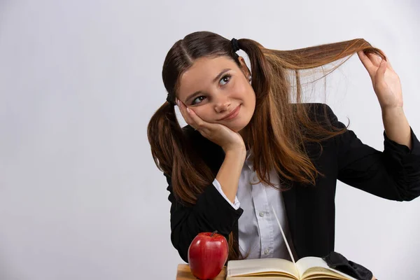 Mladá studentka si dělá domácí úkoly u stolu a rukou si drží svůj huňatý ocas. Na stole je kniha a jablko na svačinu. fotografie na šedém pozadí — Stock fotografie