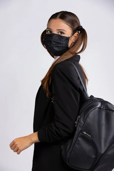 Eine junge Studentin trägt eine schwarze Gesichtsmaske, um eine Infektion mit dem Coronavirus zu verhindern, und trägt eine schwarze Jacke und einen Rucksack. Foto auf grauem Hintergrund — Stockfoto