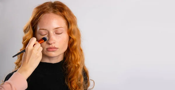 Menina com cabelo encaracolado vermelho, com maquiagem delicada, que é em pó com uma escova. Modelo elegante com aparência agradável. — Fotografia de Stock