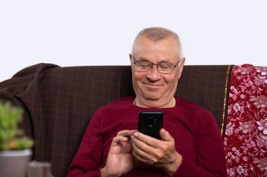 Evdeki kanepede dinlenirken elinde akıllı telefon tutan gözlüklü mutlu yaşlı bir adam. Modern teknoloji kavramı.