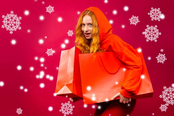 Joyeux jeune fille rousse avec des taches de rousseur tenant des sacs à provisions rouges sur un fond rouge et avec de gros flocons de neige — Photo