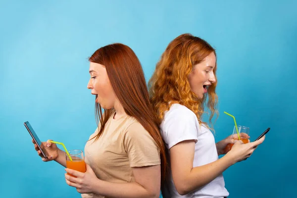 Two redhead girlfriends in casual clothes, standing back to back. Isolated on blue background. They look at their phones in surprise and drink juice. People lifestyle concept. — Stock Photo, Image