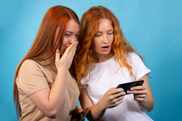 Deux copines aux cheveux roux en tenue décontractée. Isolé sur fond bleu. Je suis surpris de voir quelque chose au téléphone. Concept de style de vie. — Photo