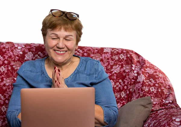 Μια όμορφη ηλικιωμένη γυναίκα, χρησιμοποιεί ένα φορητό υπολογιστή και χαμογελάει χαρούμενα σε βιντεοκλήση. Φωτογραφία σπιτιού σε καναπέ με κενό πλευρικό χώρο. — Φωτογραφία Αρχείου