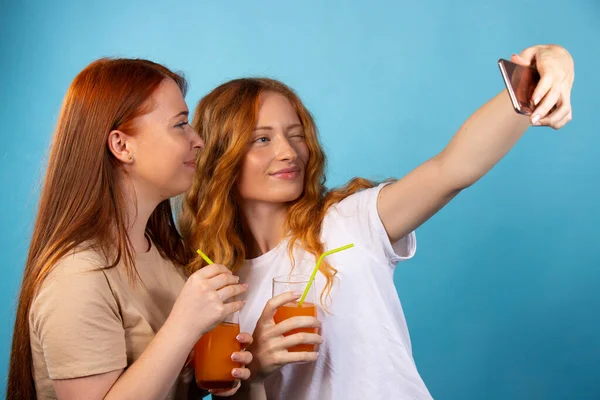 Red-haired girlfriends in casual clothes drink straw juice and take selfies. Isolated on a blue background. People lifestyle concept. — Stock Photo, Image