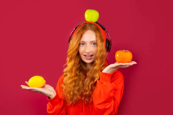 Uma linda menina ruiva com cabelos longos encaracolados com sardas, usando fones de ouvido e segurando frutas. Retrato sobre um fundo vermelho — Fotografia de Stock