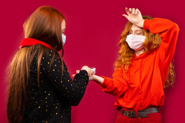 Duas meninas com máscaras médicas medem a temperatura corporal com um termômetro sem contato. Foto sobre um fundo vermelho — Fotografia de Stock