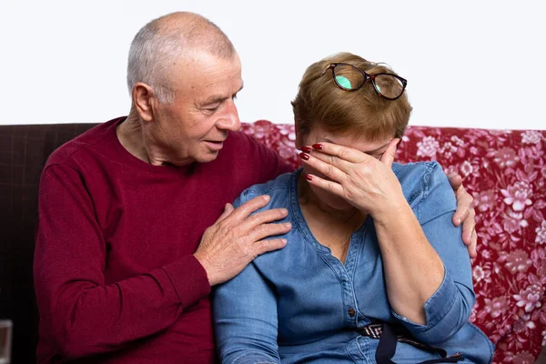 Elderly spouses are sitting on the couch at home, the wife is crying, the husband hugs her and comforts her. Sad life event