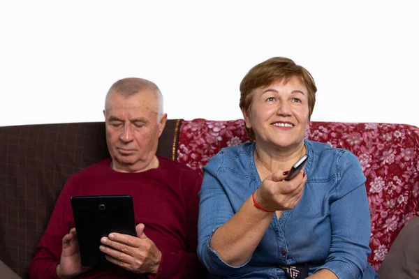 Casal sénior em casa num sofá aconchegante. Uma mulher está assistindo TV e um homem está lendo as notícias em um tablet. Conceito online sênior e conceito pandêmico. — Fotografia de Stock