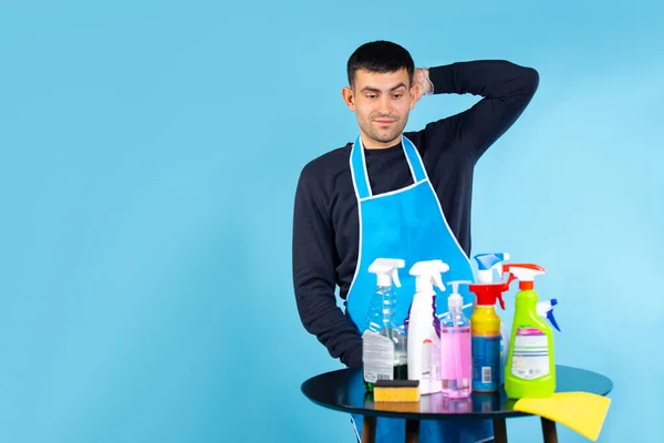 Gender stereotypes. The man holds his head and looks at the set of cleaning products. Photo on a blue background with empty side space. — Stock Photo, Image