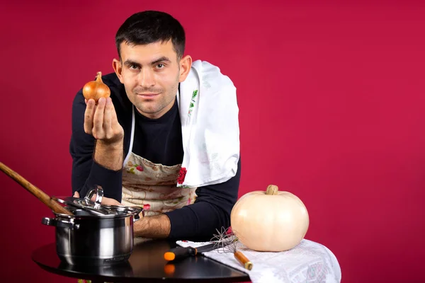 Um homem prepara comida em casa na cozinha e posa com uma cebola. Foto sobre um fundo rosa com espaço lateral vazio. O conceito de estereótipos de gênero. — Fotografia de Stock