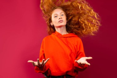 Kızıl saçlı, dalgalı kıvırcık saçlı ve kolları yanda olan bir kız. Kırmızı arka plandaki kameraya bakar. Yüksek kalite fotoğraf